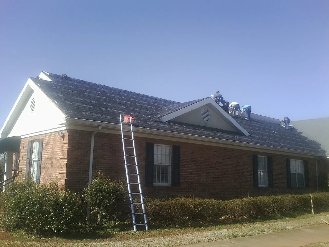 Crew tearing shingles off a brick ranch roof with a ladder set — Metal Roof Repair work by our crew in Pembroke Township