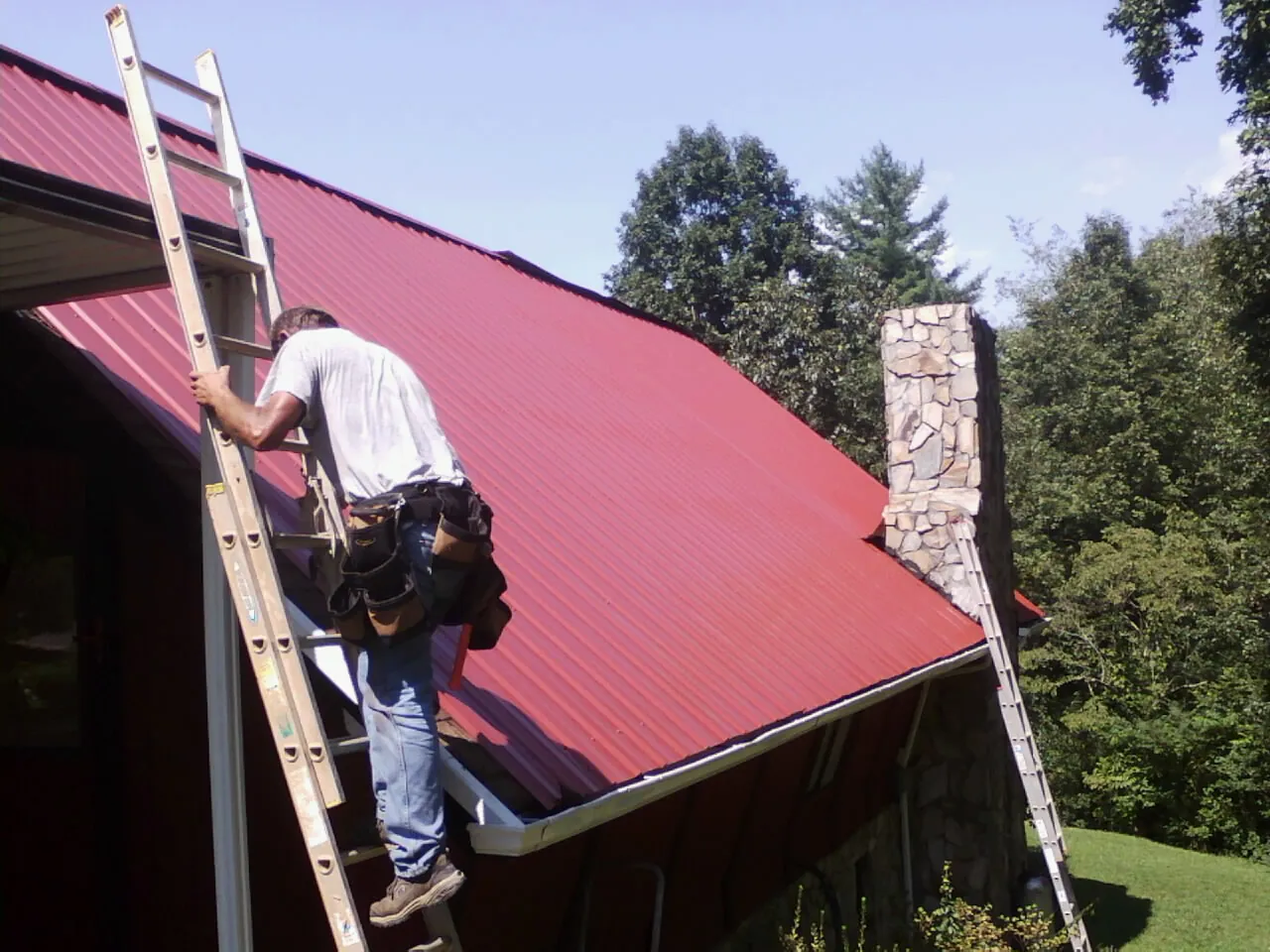 Roofer on a ladder beside a red metal roof and stone chimney — Soffit & Fascia Repair by Roofing Brothers in Pembroke Township